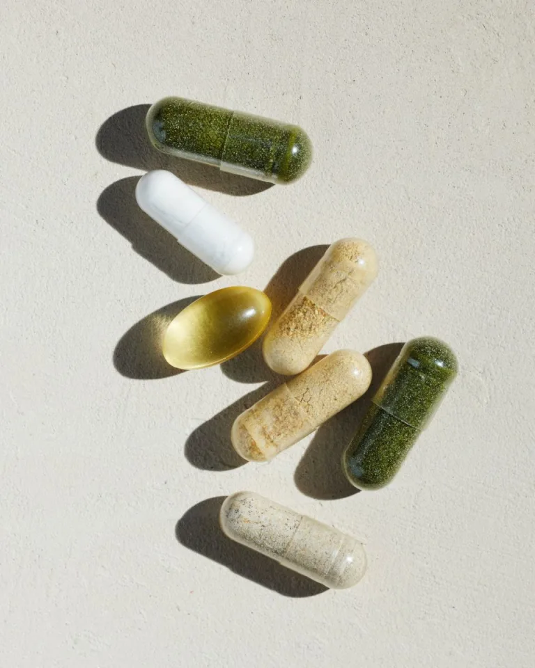 Sakara The Foundation: Prenatal assorted supplements including green and beige capsules, white capsule, and golden omega softgel shown on a textured light surface in directional sunlight.