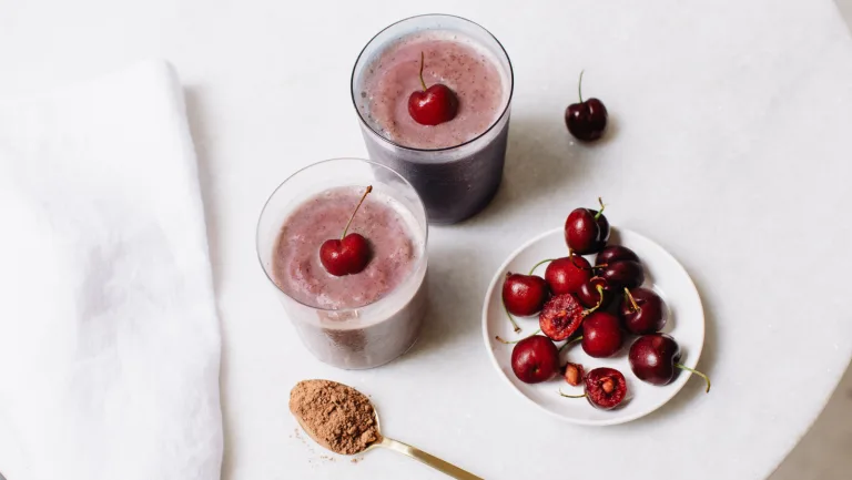 Chocolate-Cherry Metabolism Super Smoothies in glasses topped with whole cherries, with fresh cherries and Metabolism Super Powder nearby in a spoon.