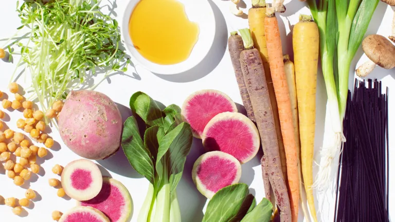 Overhead arrangement of fresh vegetables including carrots, watermelon radishes, leafy greens, chickpeas, and oil in a small dish.