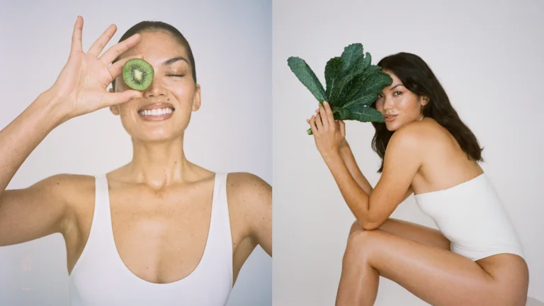Woman holding a kiwi slice over one eye beside another woman holding leafy greens.