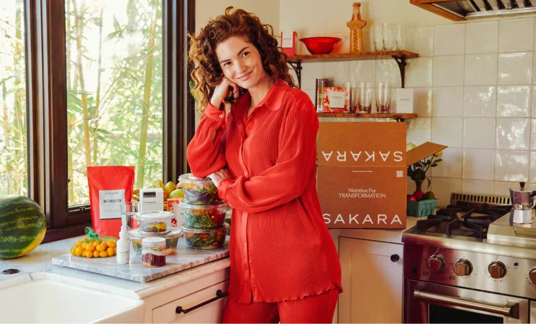 Woman in a red outfit standing in a kitchen beside stacked prepared Sakara meal containers and a brown delivery box labeled Sakara.