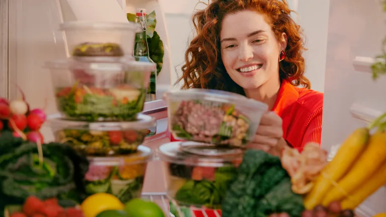 Woman smiling while placing clear Sakara containers of prepared salads into a refrigerator filled with fresh produce.
