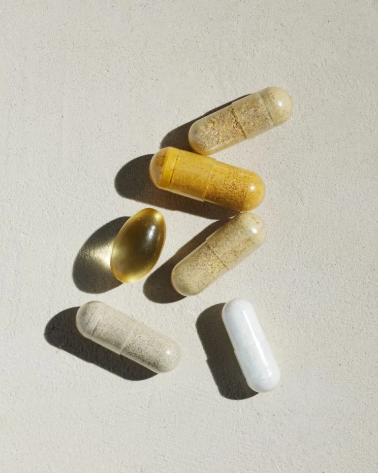 Sakara The Foundation assorted capsules and softgels shown close-up on a textured light surface with soft directional sunlight casting shadows.