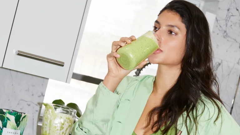 Woman in a kitchen drinking a green smoothie from a glass.
