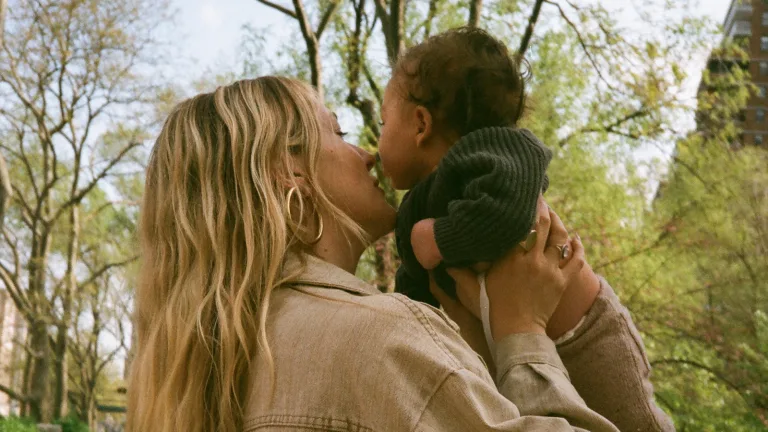 Woman lifting and kissing a baby outdoors in a park setting.