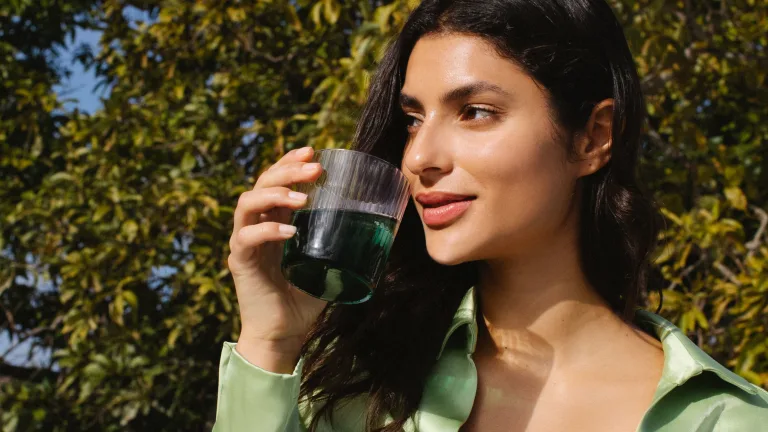 Woman outdoors holding a glass of water mixed with chlorophyll against a backdrop of leafy trees.