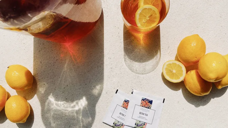 Overhead view of lemon, a glass of tea with lemon, a pitcher of tea, and packets of Sakara Detox Tea on a light surface.