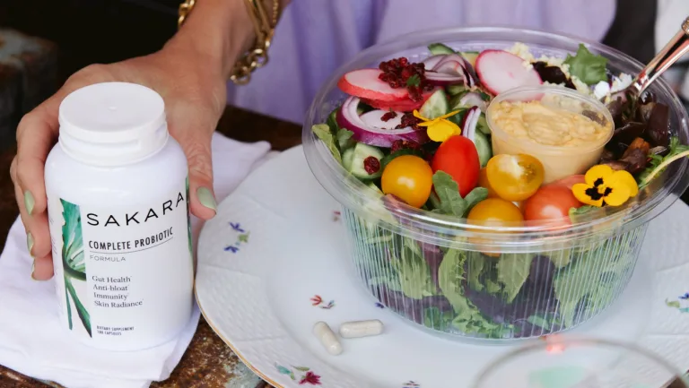 A container of Sakara salad next to a hand holding sakara.momplete Probiotic.