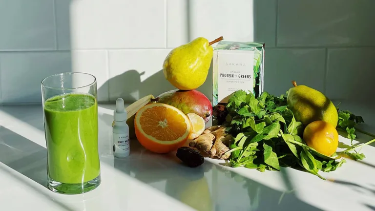Glass of green smoothie beside fresh citrus, leafy greens, ginger, and a box of Protein + Greens on a tiled surface.
