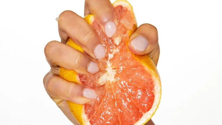 Hand squeezing a halved grapefruit with juice dripping from the fruit against a light background.