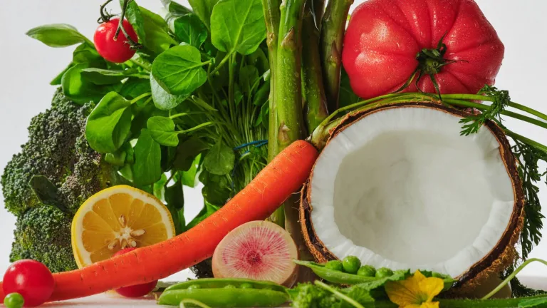 Assorted fresh produce including tomatoes, carrots, citrus, coconut, and leafy greens arranged against a white backdrop.