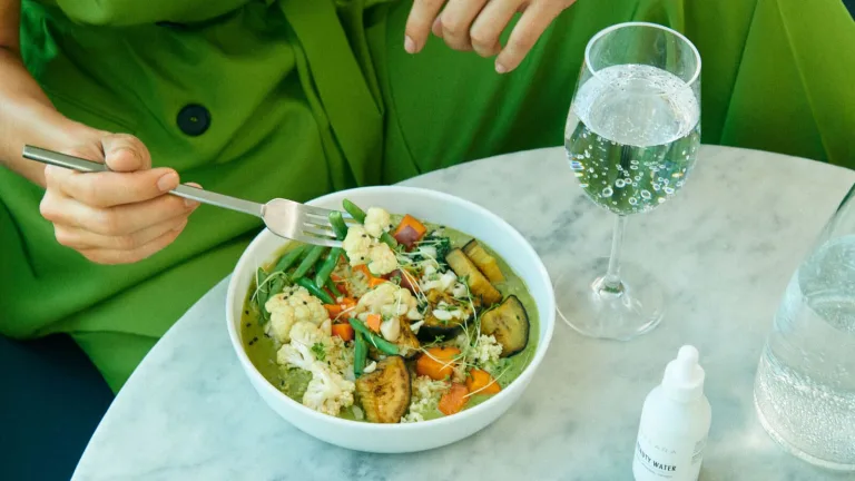 Person in a bright green blazer eating a bowl of Sakara curry at a marble table with a glass of sparkling water.