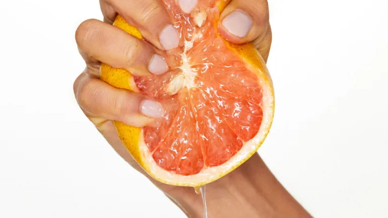 Hand squeezing a halved grapefruit with juice dripping from the fruit against a light background.