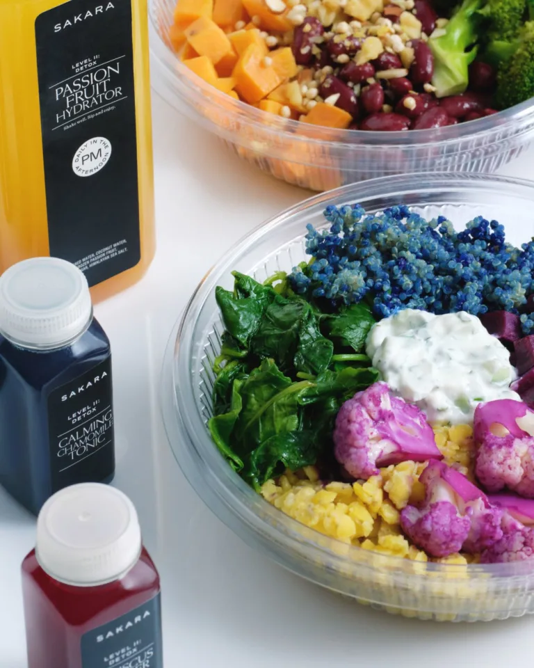 Sakara Level II: Detox ready-to-eat meal in a clear round container with leafy greens, blue quinoa, yellow grains, pink cauliflower, beets, and creamy dressing, displayed beside bottled tonics and Passion Fruit Hydrator.
