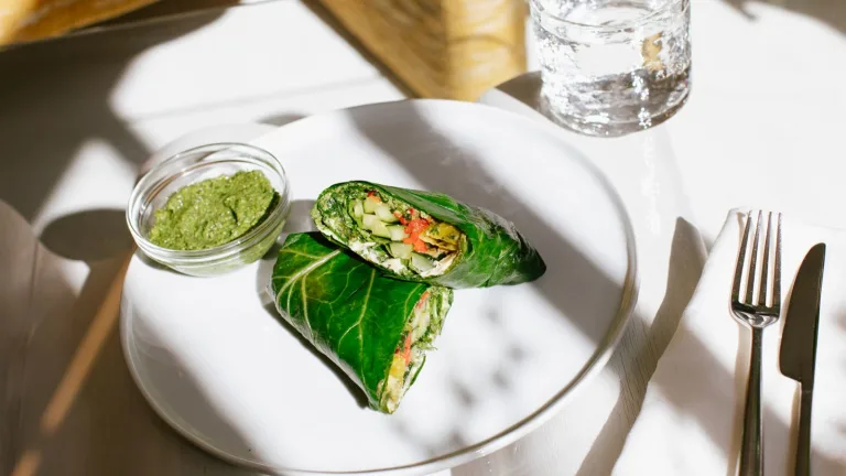 Collard green wrap sliced in half on a white plate with a small bowl of green dipping sauce.