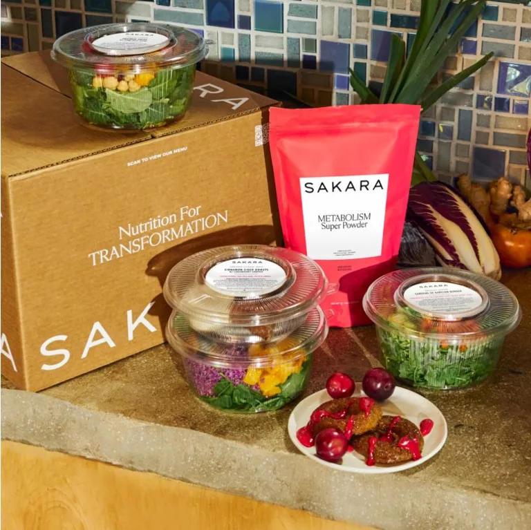 Sakara Signature Nutrition Program packaging featuring a brown Sakara delivery box labeled “Nutrition For Transformation,” clear ready-to-eat breakfast and lunch containers, and a red Metabolism Super Powder pouch displayed on a kitchen counter.