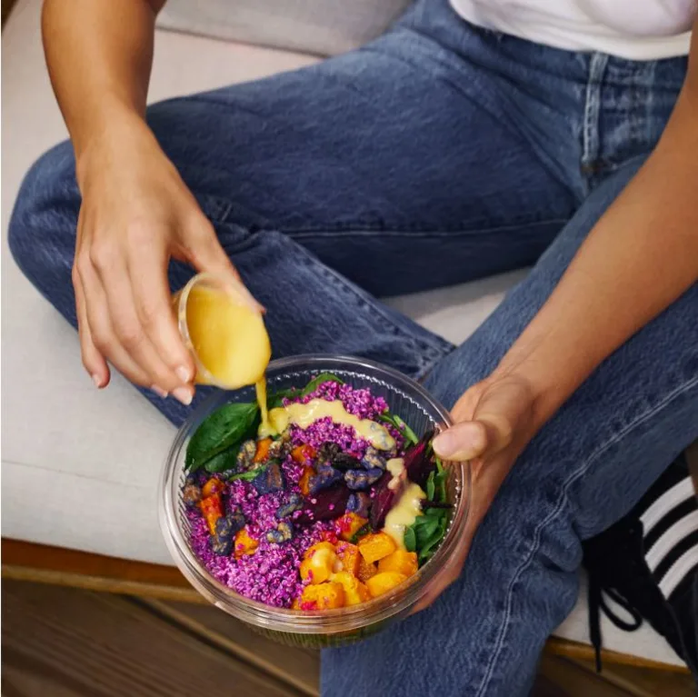 Sakara Signature Nutrition Program ready-to-eat plant-based meal in a clear round container topped with leafy greens, roasted vegetables, grains, and dressing being poured over the bowl.