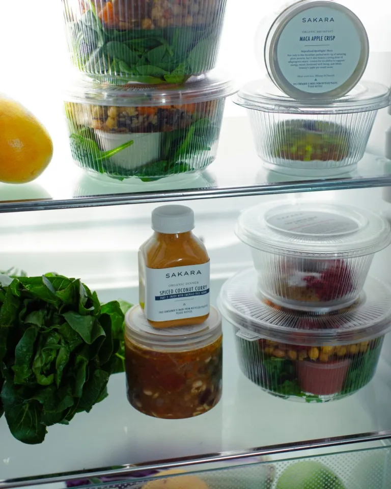 Sakara Signature Nutrition Program packaging labeled breakfast, lunch, and dinner in clear ready-to-eat containers and bottled Spiced Coconut Curry displayed inside a refrigerator beside fresh leafy greens and citrus produce.
