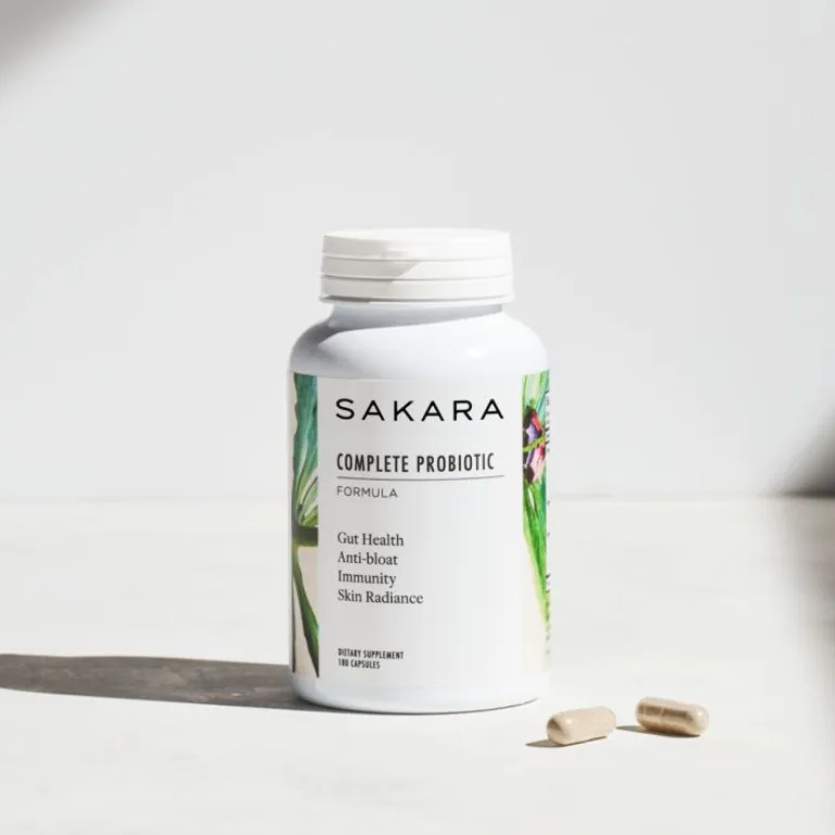 sakara.momplete Probiotic Formula in white bottle with white cap, 90 capsules, labeled gut health, anti-bloat, immunity, and skin radiance, shown with two capsules on surface.