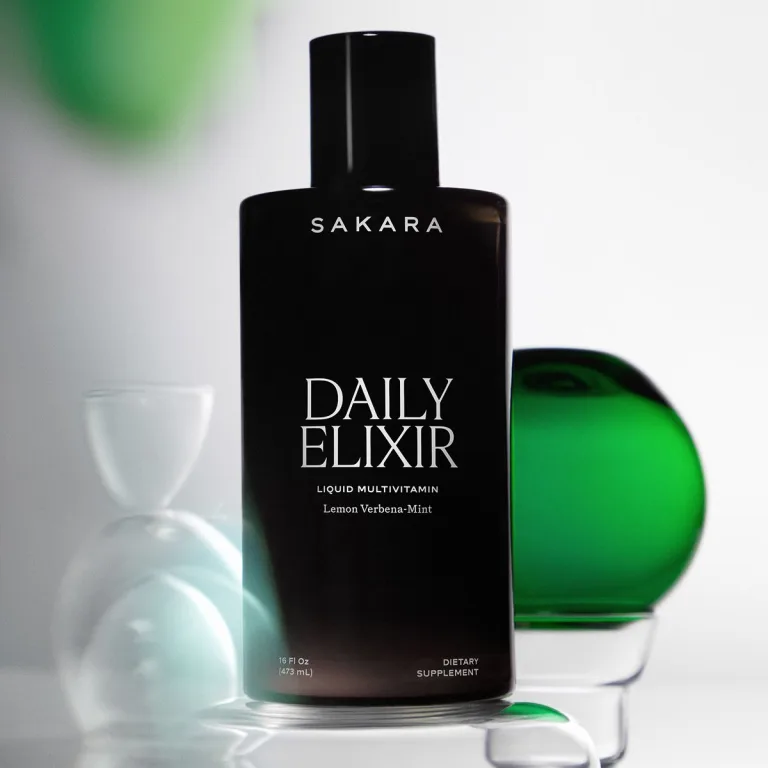 Sakara Daily Elixir liquid multivitamin in matte black 16 fl oz bottle labeled Lemon Verbena-Mint on neutral background with green glass accent and white sculptural objects