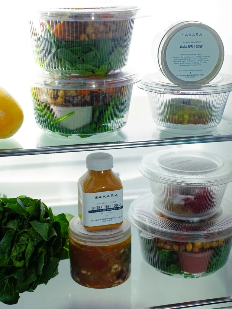 Sakara Signature Nutrition Program refrigerated meals in clear ready-to-eat containers displayed on glass shelves beside leafy greens and citrus.