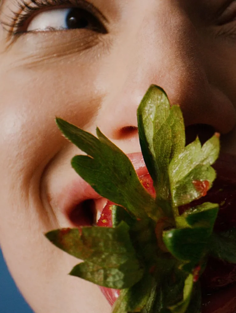 Close-up of a person holding fresh strawberries with green leaves near their mouth against a blue background.