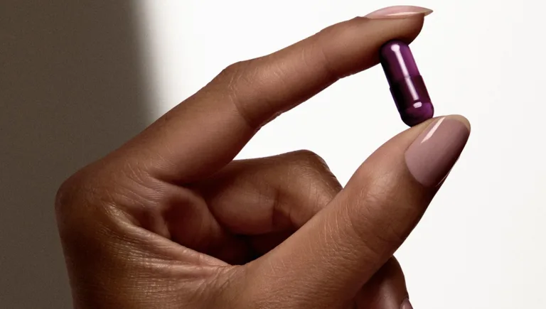 Close-up of a hand holding a small purple capsule (Sakara Night Service) between fingers against a light background.