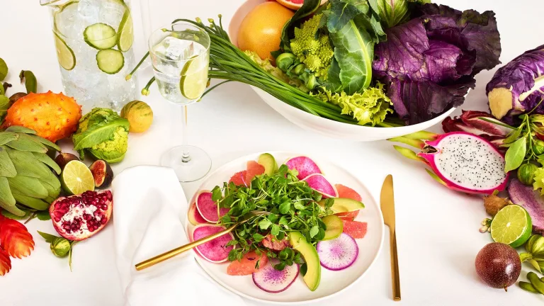 Overhead view of a colorful salad surrounded by fresh produce including citrus, artichokes, radishes, and leafy greens on a white table