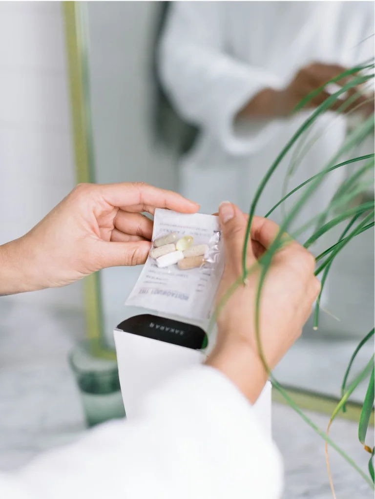 Sakara The Foundation daily supplement sachet being removed from box by hand near a plant in a bright indoor setting.