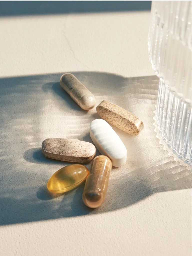 Sakara The Foundation assorted capsules and golden softgel shown close-up on a textured surface with a glass of water casting light reflections.