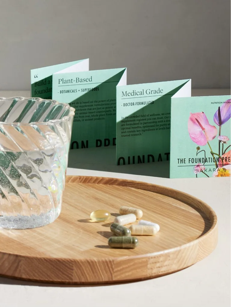 Sakara The Foundation: Prenatal assorted capsules and softgels displayed on a wooden tray beside a glass of water with mint green product booklet in the background.