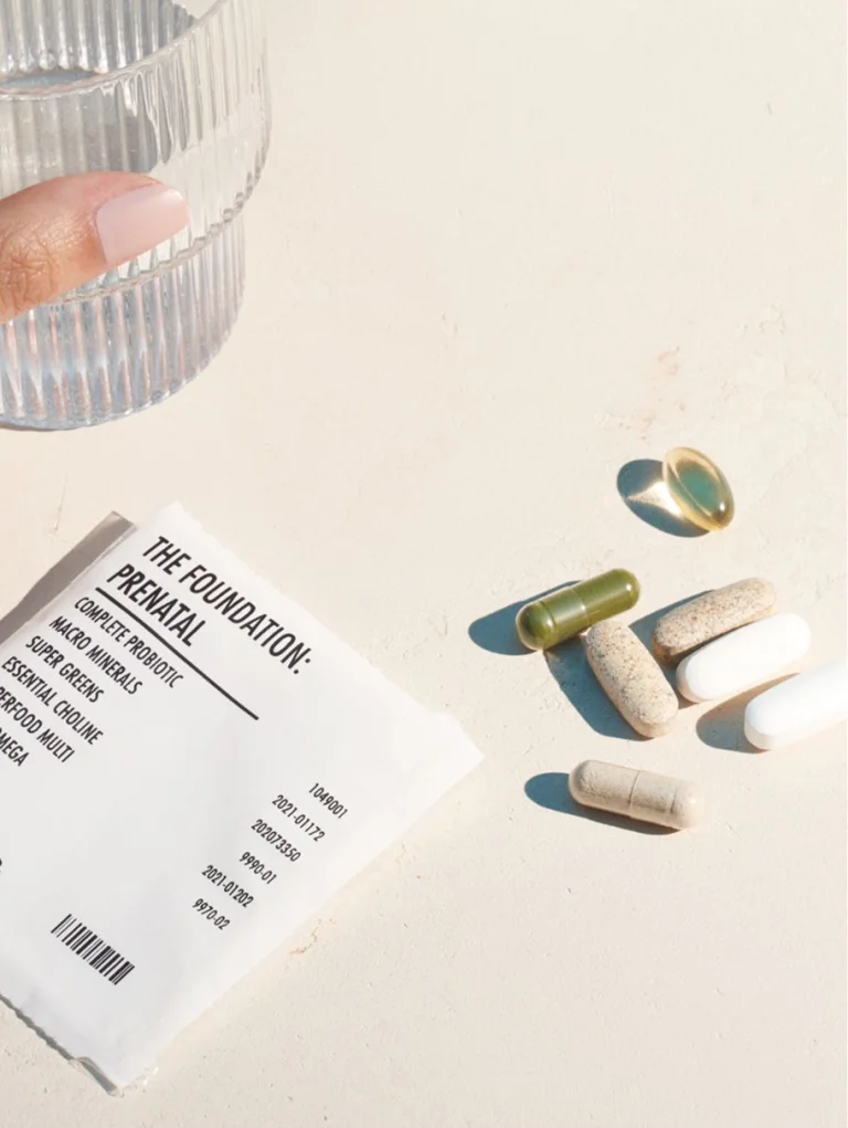 Sakara The Foundation: Prenatal daily supplement sachet shown with assorted capsules and softgels on a light textured surface beside a glass of water.