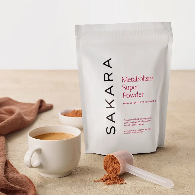 Sakara Metabolism Super Powder in matte white resealable pouch, dark chocolate flavored, 9.7 oz (276 g), shown with coffee cup and scoop of powder on neutral surface.