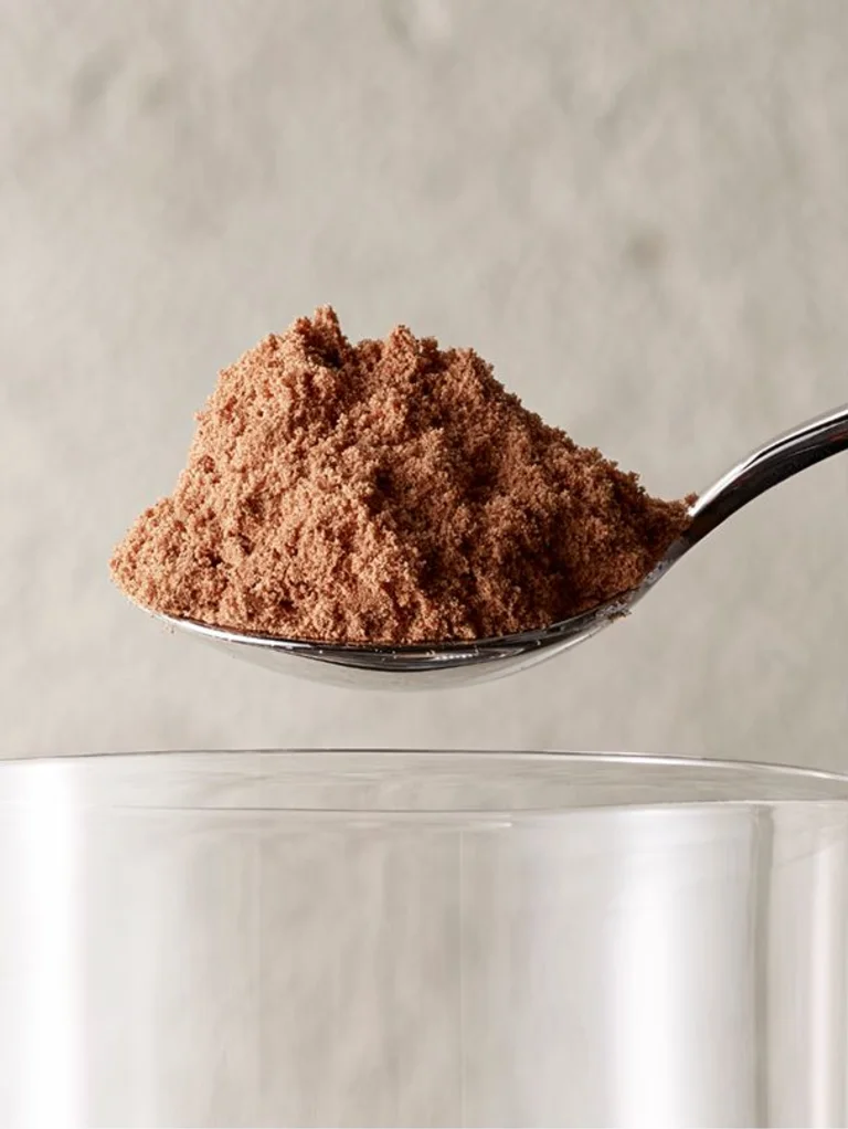 Sakara Metabolism Super Powder dark chocolate flavored powder shown heaped on a spoon above a glass of water, demonstrating stirring it into water.