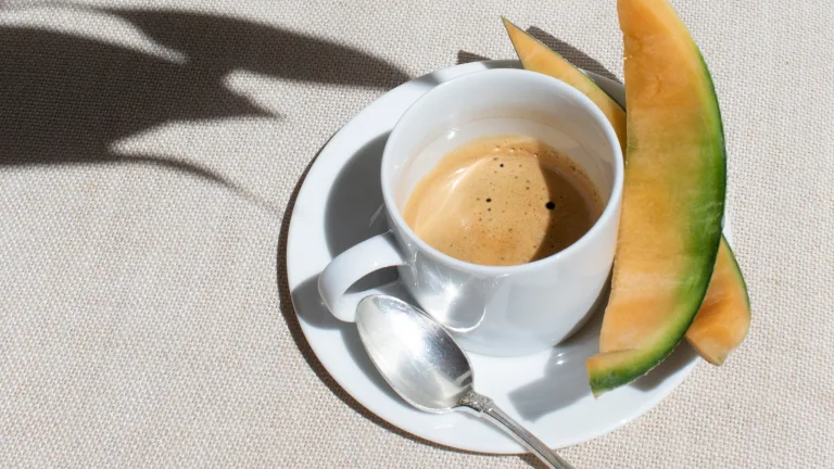 Cup of espresso on a saucer with a spoon beside slices of cantaloupe on a textured surface.